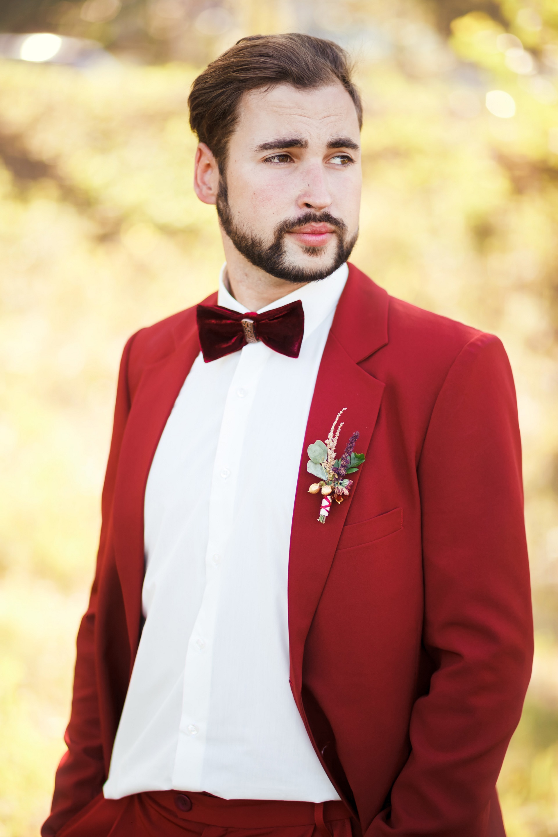 elegancki Pan Młody z brodą w czerwonym garniturze z bordową muchą i butonierką z trawami w stylu boho