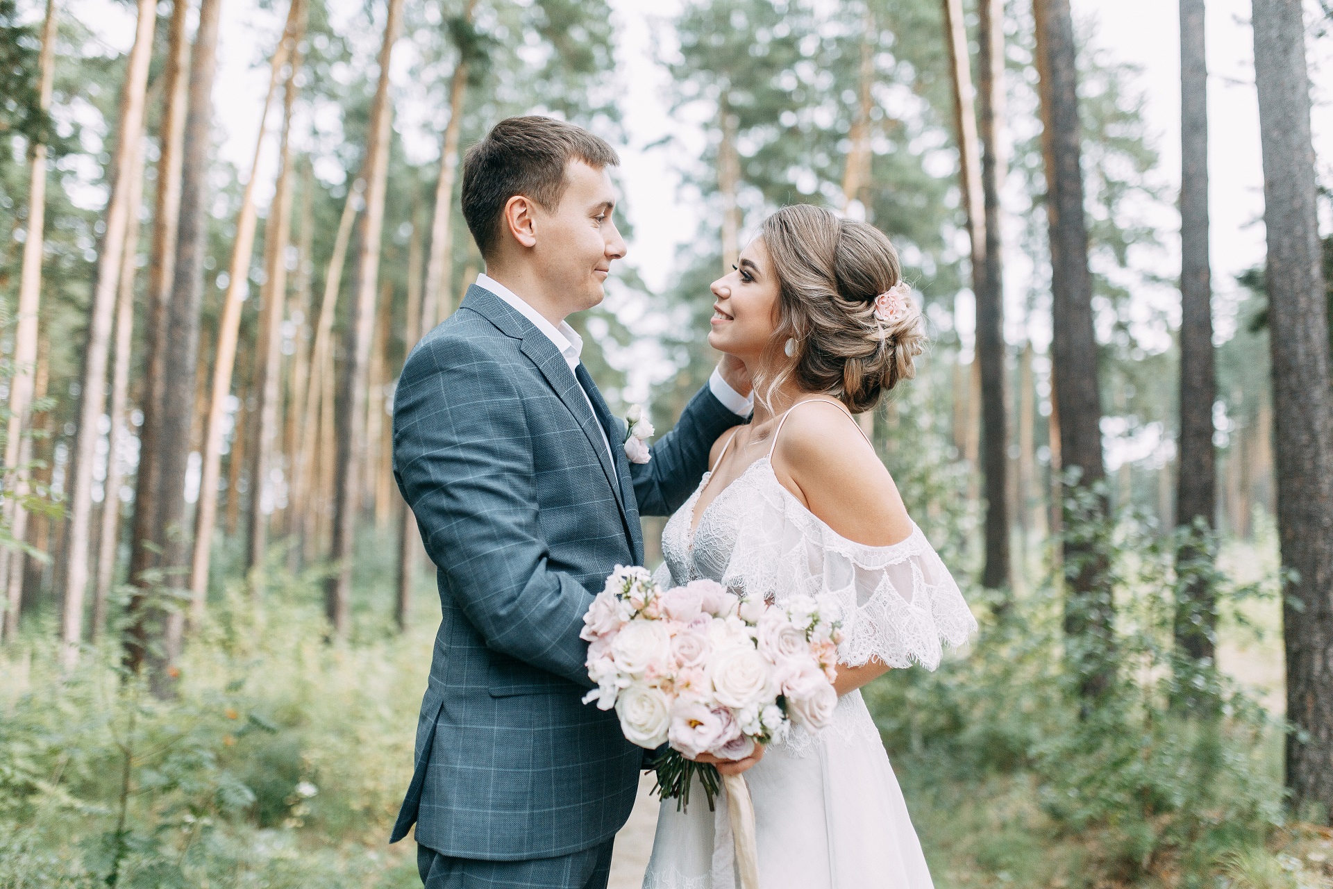 Sesja plenerowa w lesie, niebieski garnitur w kratę Pana Młodego, Panna Młoda w sukni ślubnej z opadającymi rękawkami z pastelowym bukietem ślubnym