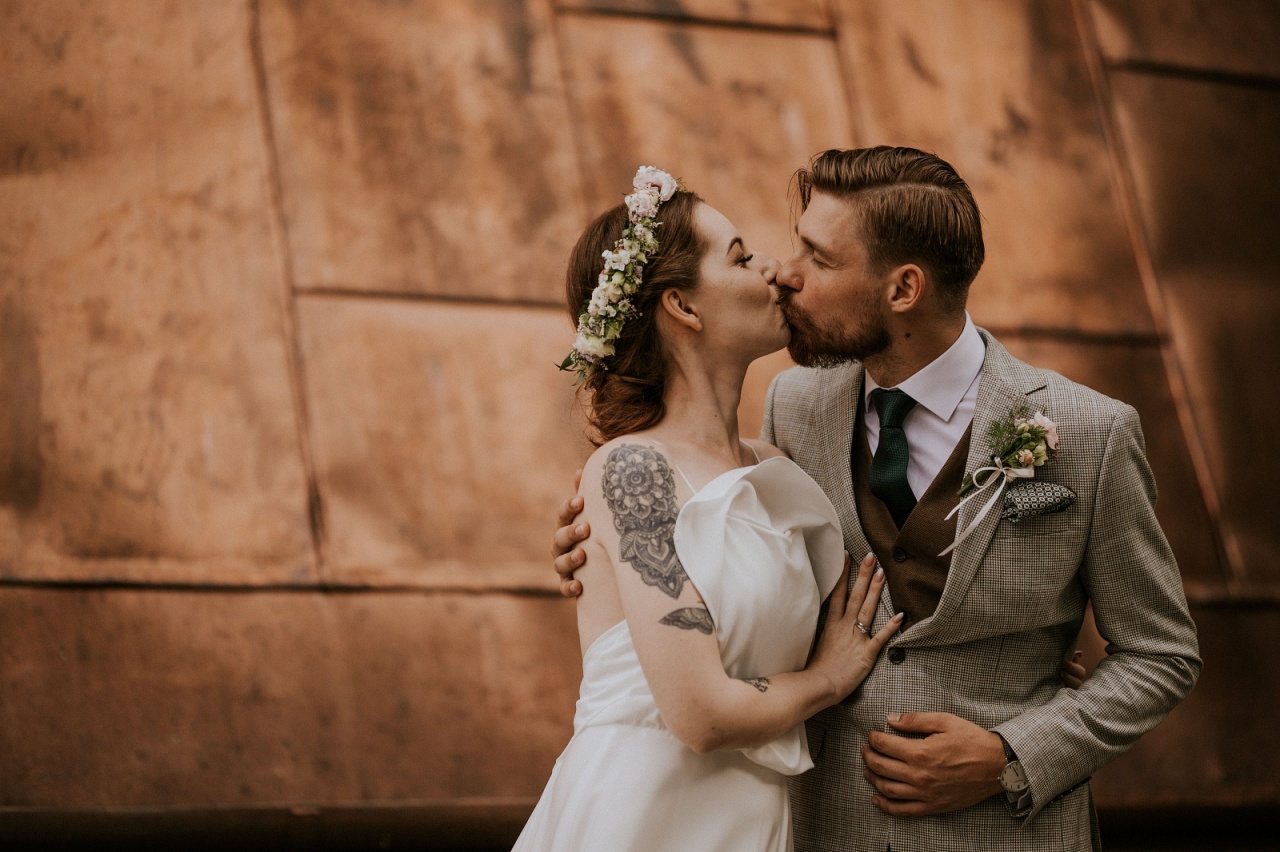 Całująca się Para Młoda, Pan Młody w jasnej marynarce z jasną butonierką i elegancką fryzurą ślubną, Panna Młoda z wiankiem na włosach i tatuażem na ramieniu