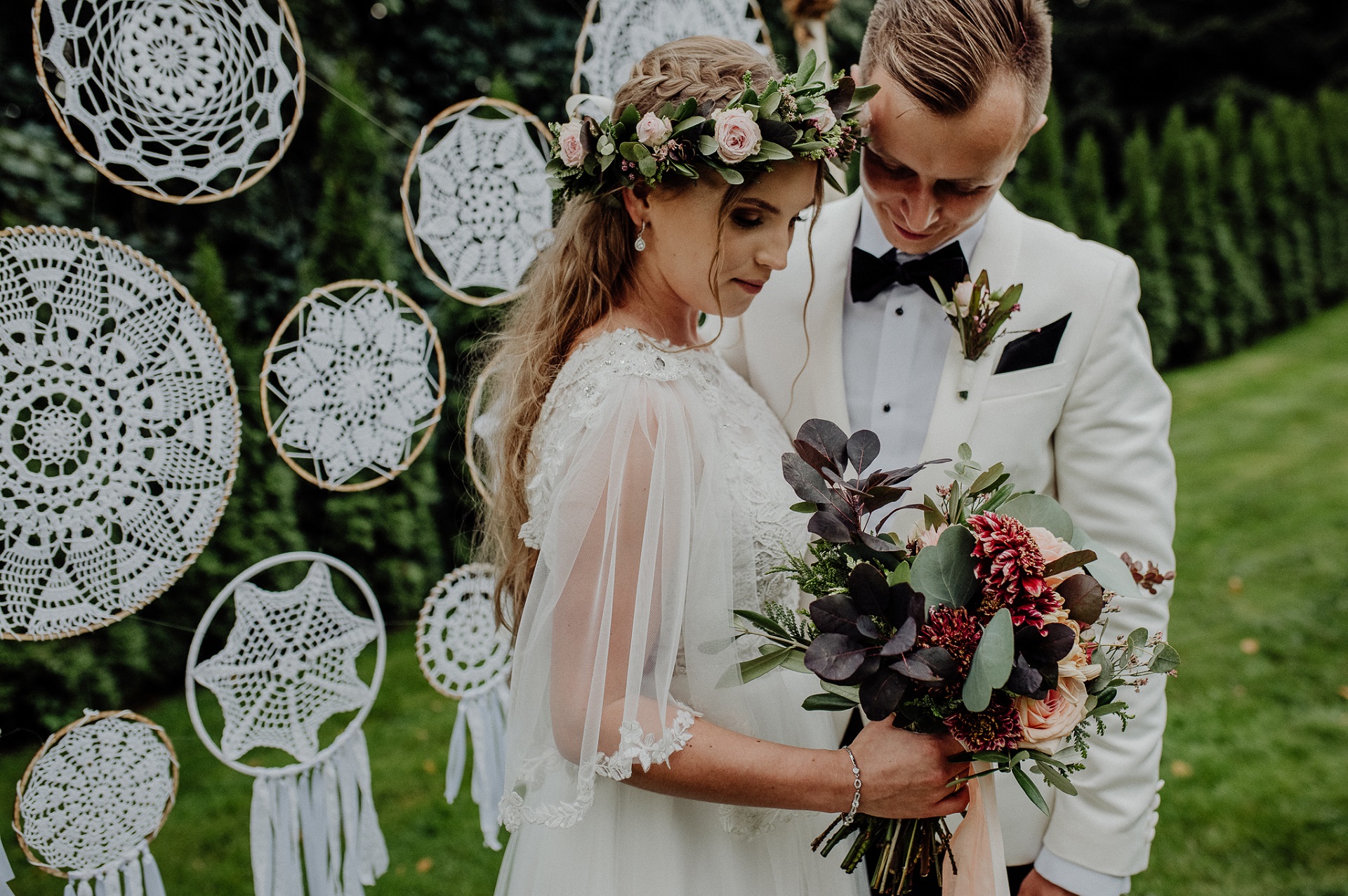 Para Młoda w stylu rustykalnym na tle dekoracji z w stylu boho. Panna Młoda w zielonym wianku z długimi włosami trzyma bukiet ślubny z ciemnych kwiatów, Pan Młody w białym garniturze z zieloną butonierką.