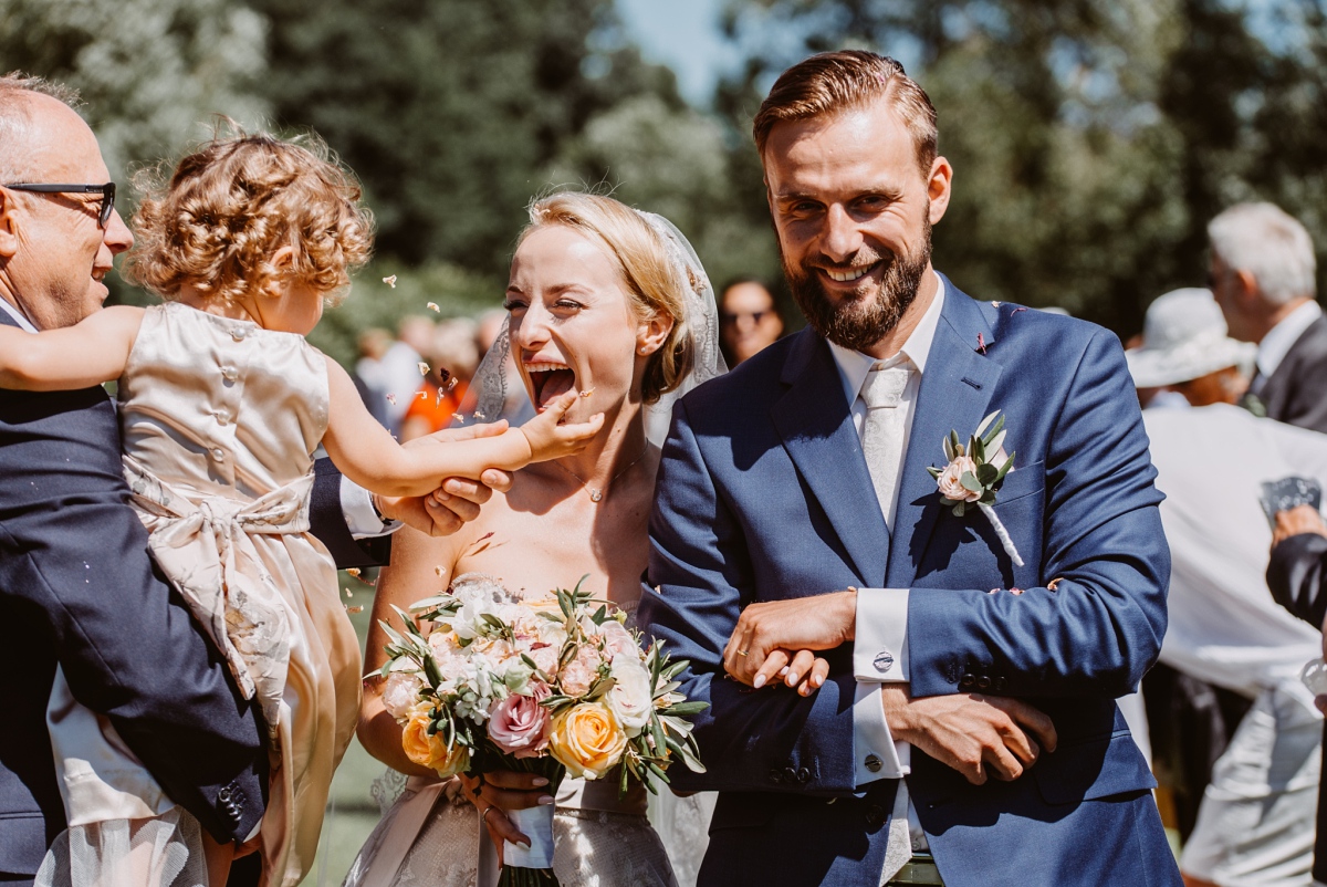 Para Młoda podczas reportażu ślubnego w ogrodzie, Pan Młody w jasnym niebieskim garniturze z pastelową butonierką, Panna Młoda trzyma pastelowy bukiet ślubny