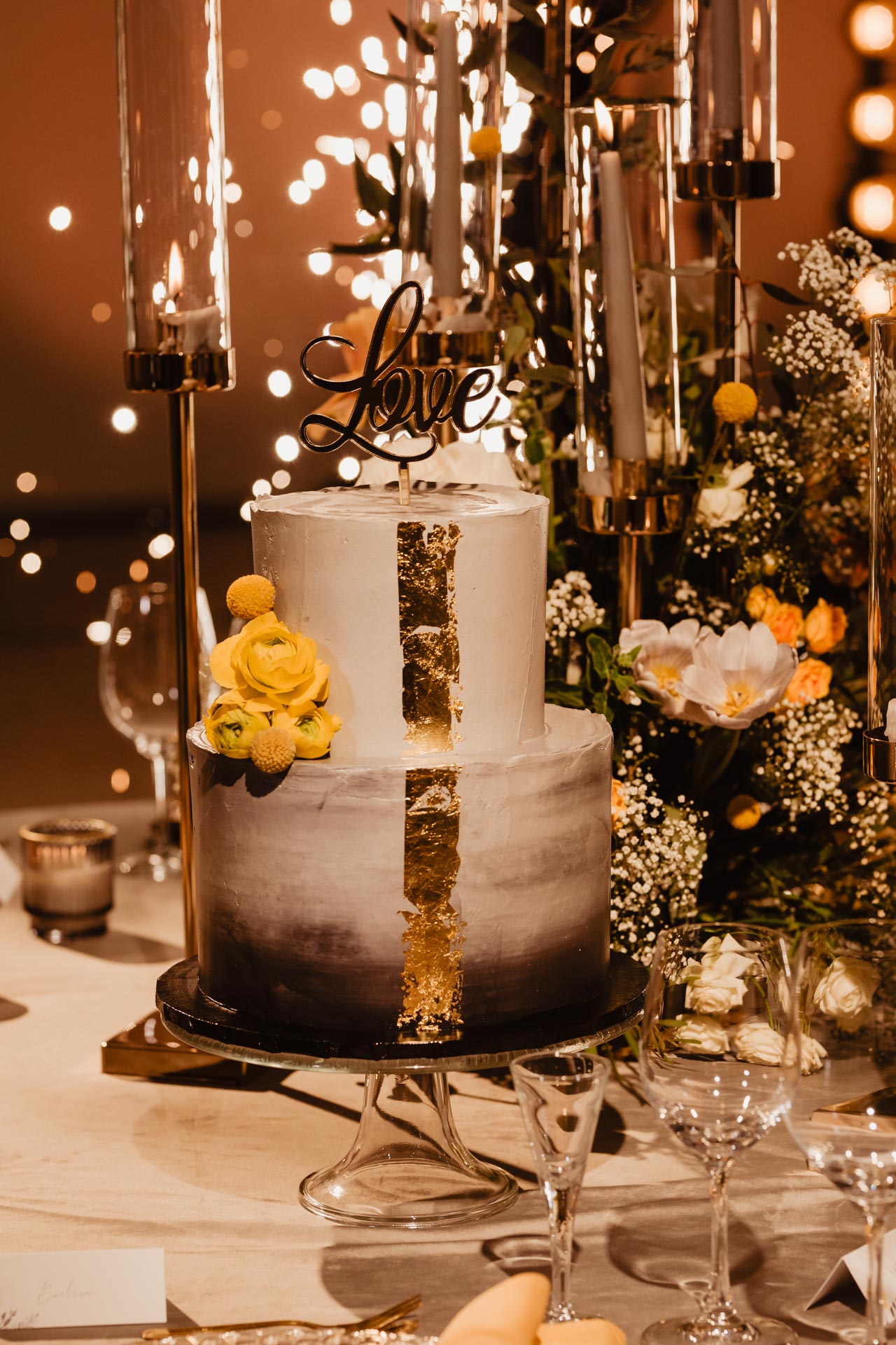 Szary tort weselny typu naked cake ze złotym topperem i złotymi zdobieniami na stole podczas wesela